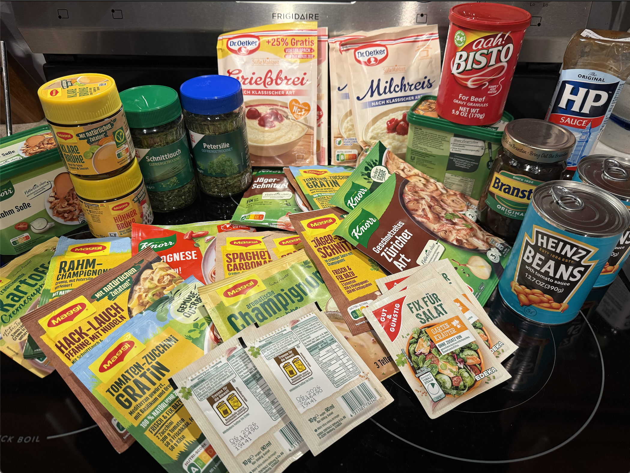 Nigel's actual pantry shelf: Maggi fix packets, Knorr Rahm sauce, EDEKA salad dressing packets, and British stock like HP Sauce, Branston pickle, and Heinz beans